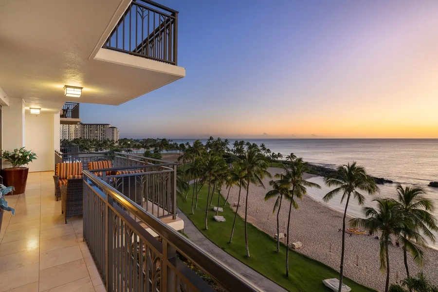 A balcony overlooks palm trees, a beach, and the ocean at sunset.