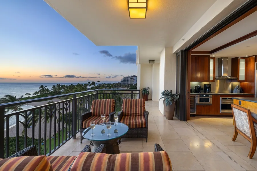 A furnished balcony overlooks the ocean at sunset, with a kitchen visible through open doors.