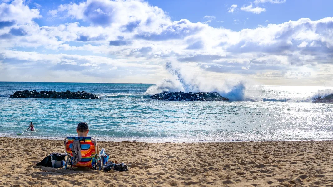 A person sits on a sandy beach watching waves break against a rocky shoreline.