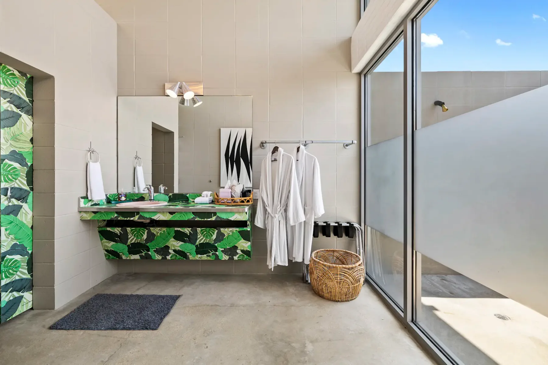 Modern bathroom with green leaf-patterned tile, double sink, two white robes, basket, towel racks, and a large window.