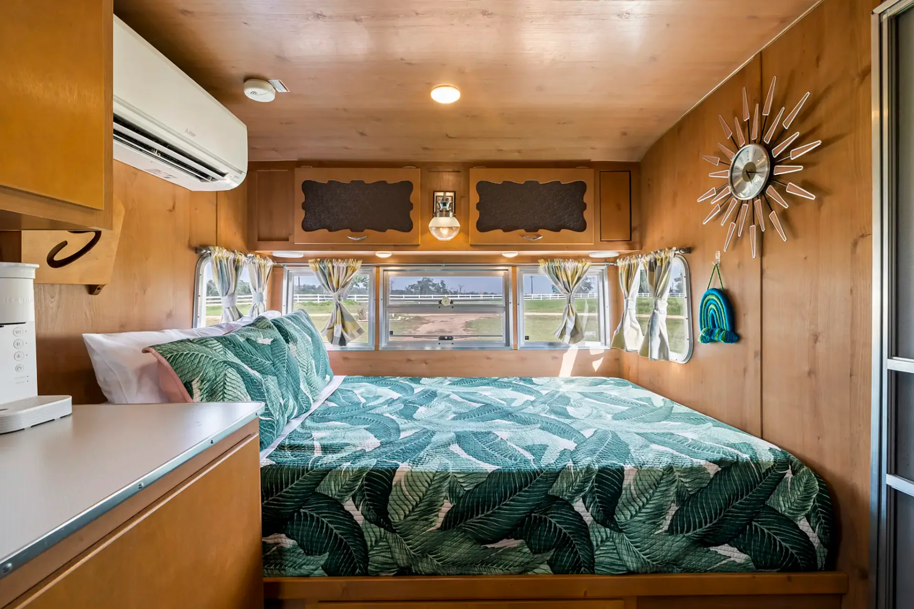Cozy trailer bedroom with a leafy green bedspread, wood paneling, sun-shaped wall clock, and a large window showing a scenic outdoor view.