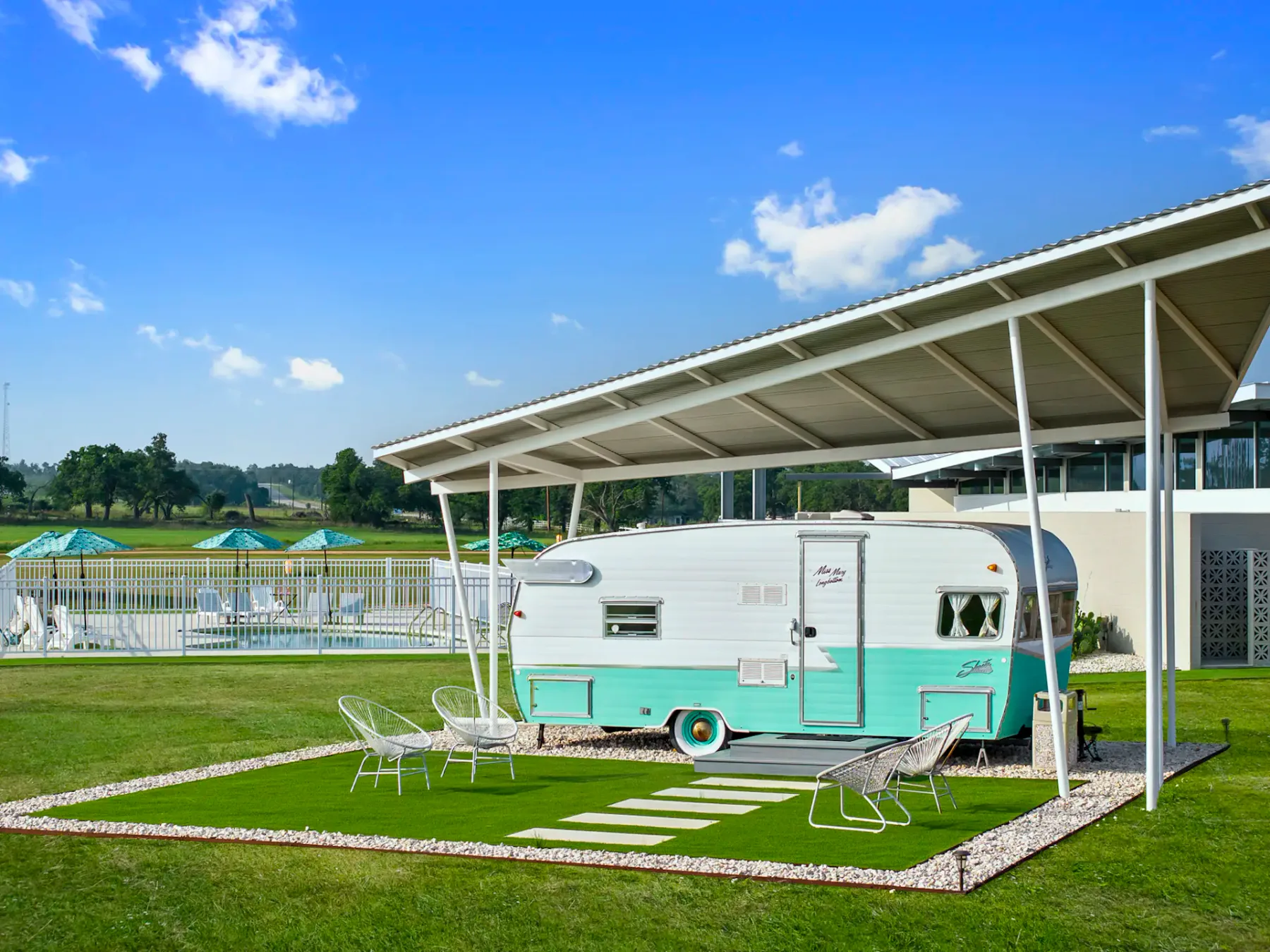 A vintage turquoise and white camper under a metal canopy with patio chairs on a grass area, set beside a pool and lush landscape.