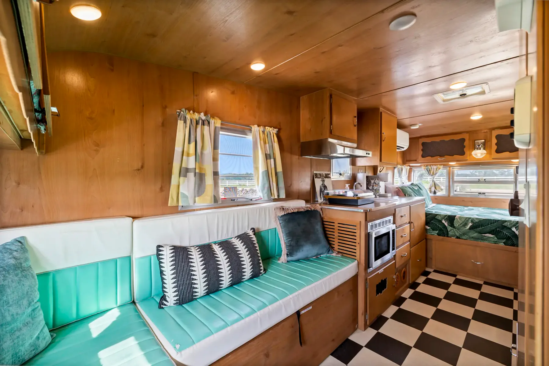 Interior of a cozy camper van with wooden paneling, teal cushioned seating, a small kitchen, chequered floor, and a bed in the back.