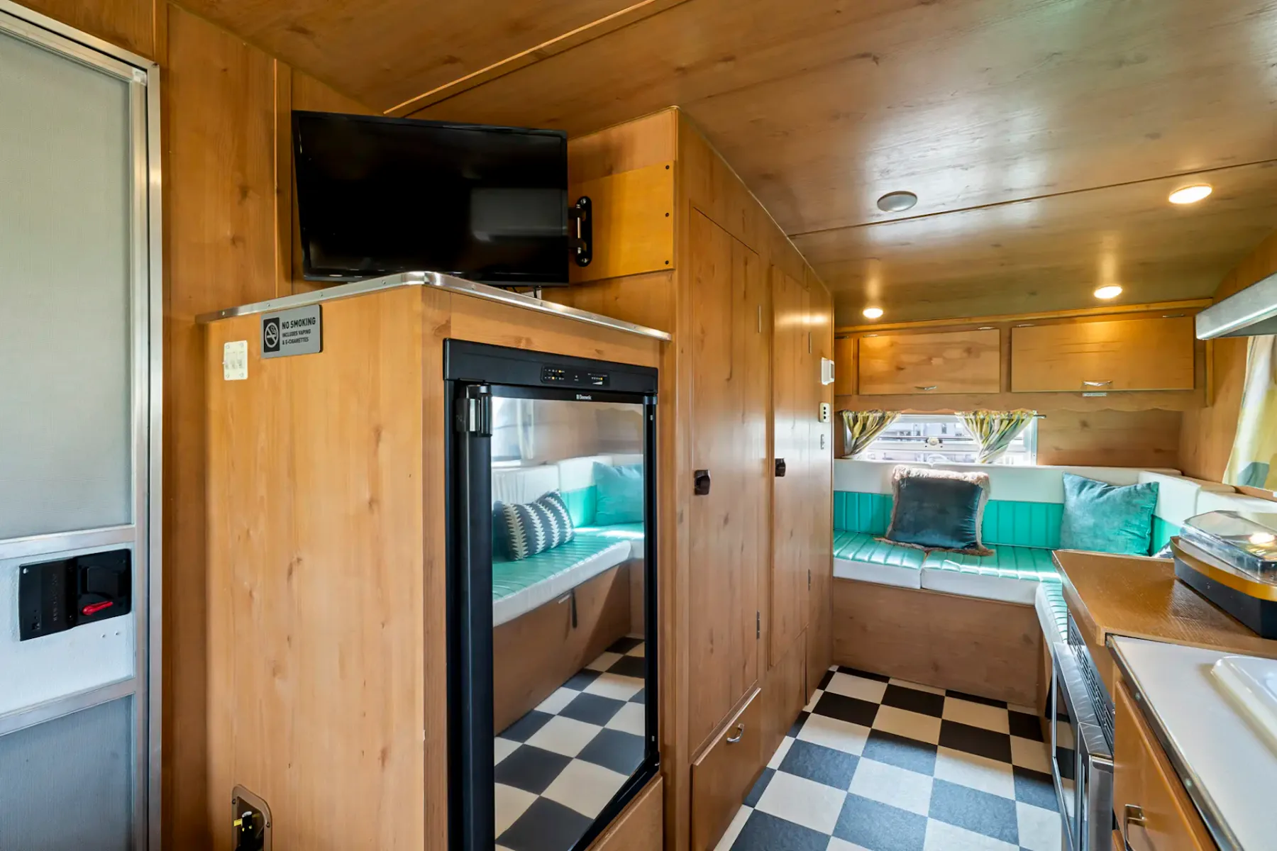 Interior of a cozy camper van with wooden cabinets, checkered floor, and a seating area. Includes a sink, refrigerator, and small TV.
