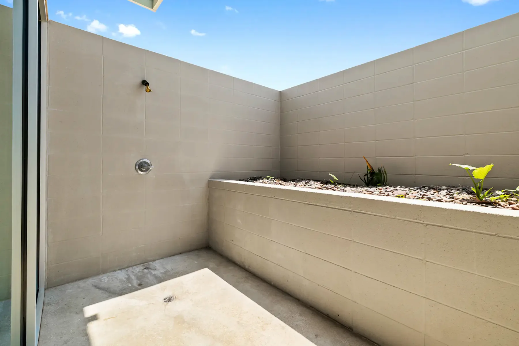 Enclosed concrete patio with a minimalist design, featuring a small raised planter box with a few plants and a blue sky overhead.