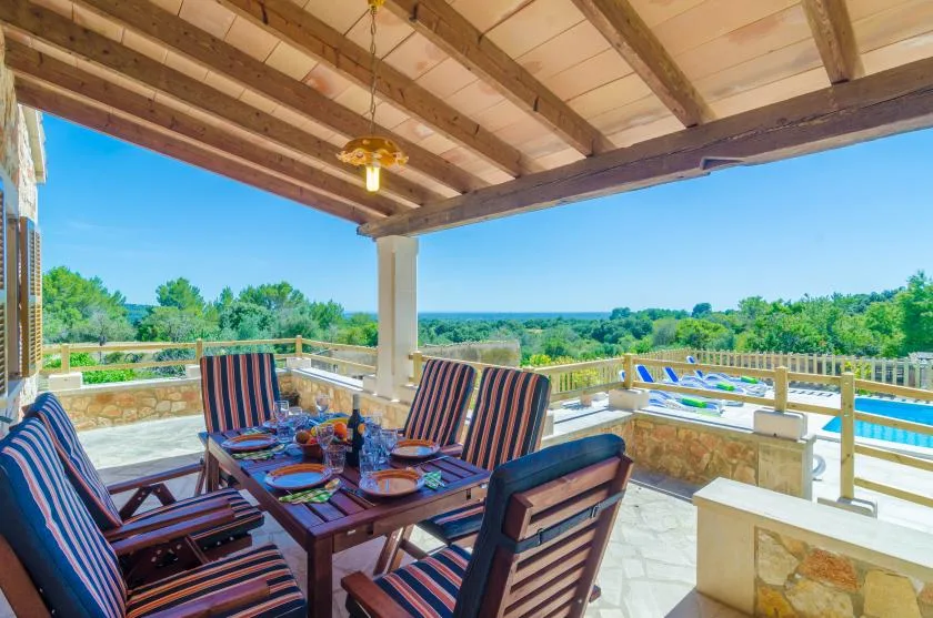 Un patio cubierto tiene asientos a rayas alrededor de una mesa, con vistas a una piscina y un paisaje verde.