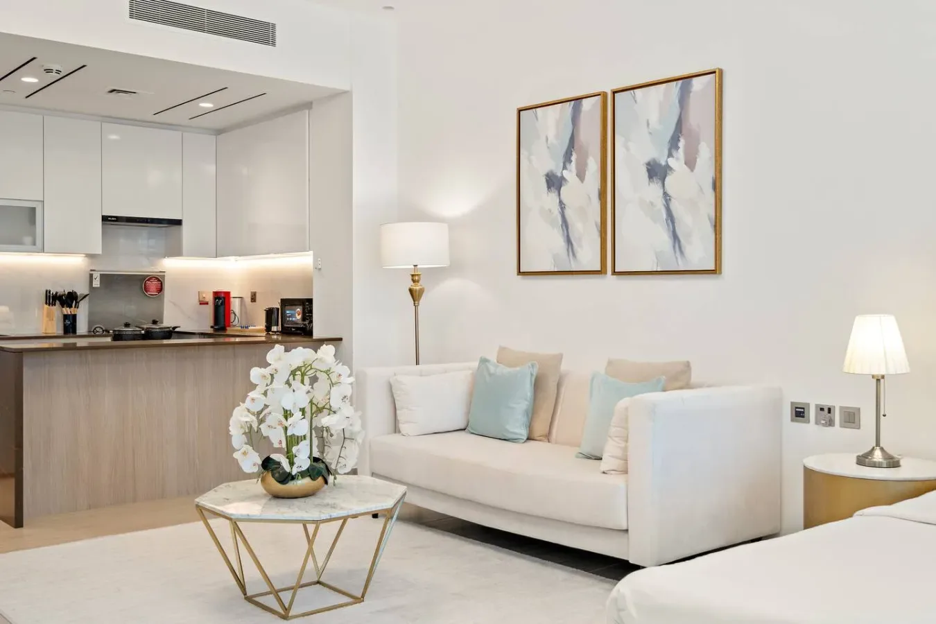 Una sala luminosa con asientos claros, cuadros abstractos y una cocina abierta.