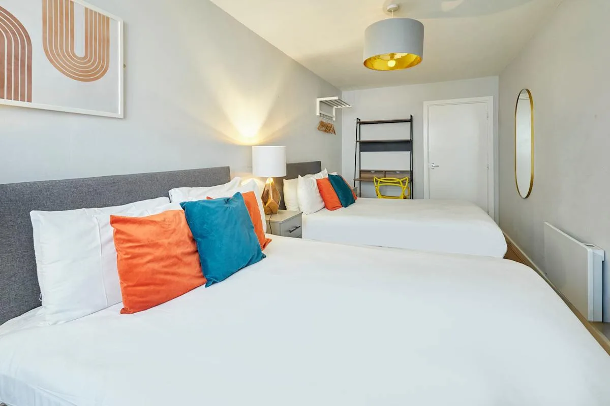 A bedroom with two beds, colorful pillows, and a bedside lamp.