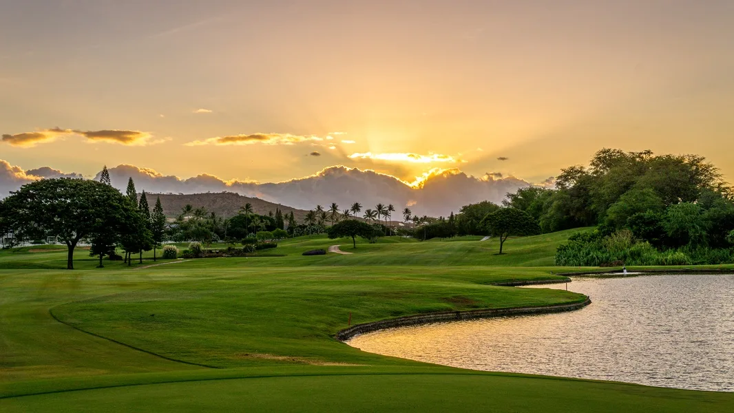 Un campo de golf se extiende junto a un estanque bajo un cielo resplandeciente al atardecer.