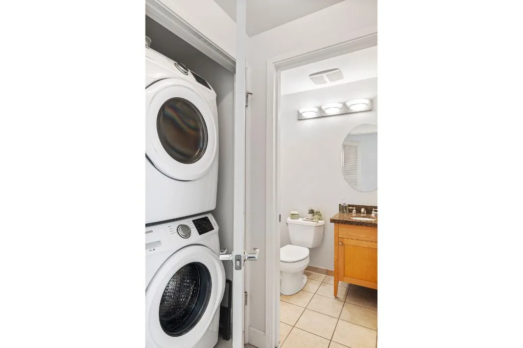 Un baño con lavabo e inodoro, visto a través de una puerta abierta junto a una lavadora y secadora apiladas.