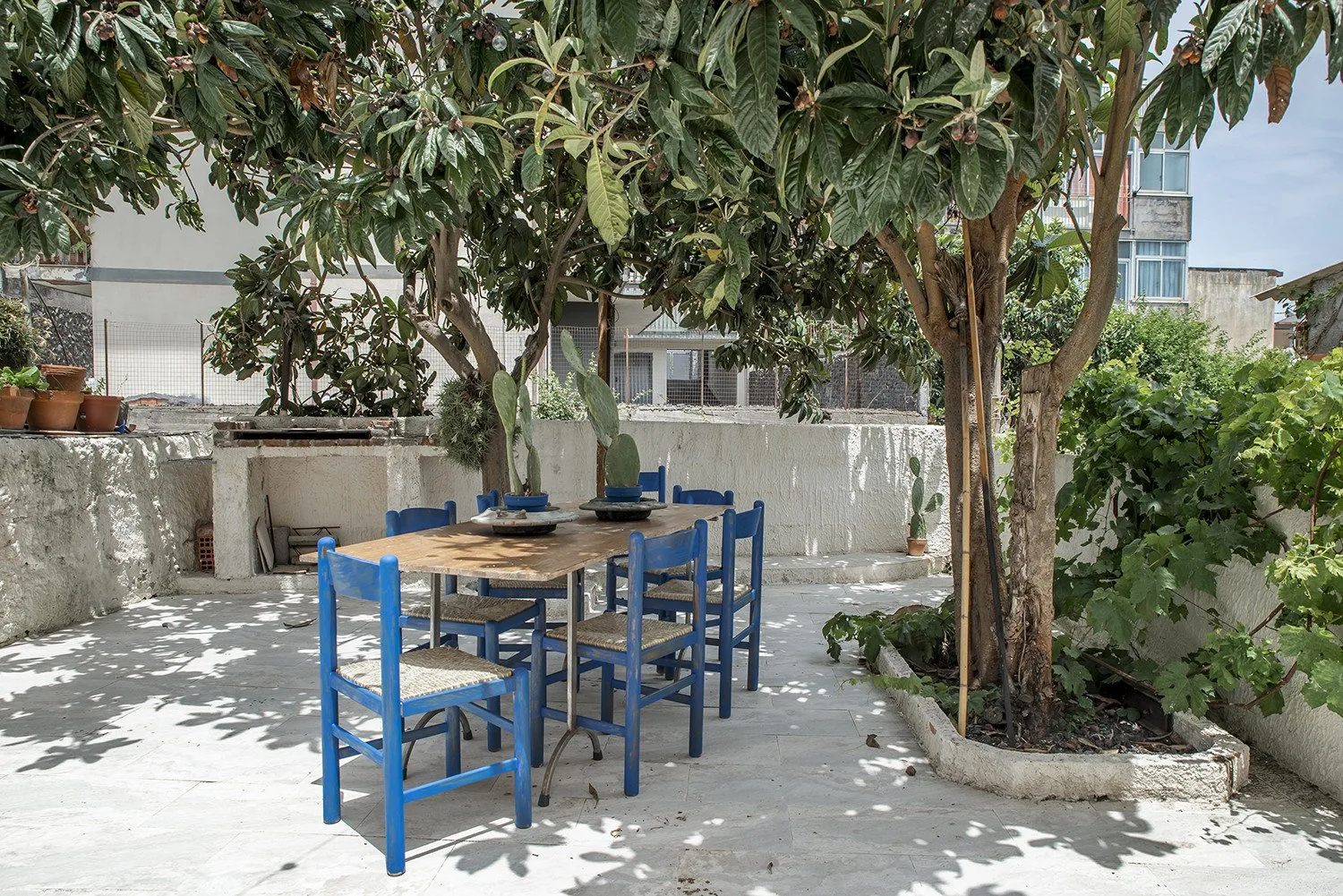 Una zona de comedor al aire libre tiene una mesa de madera y asientos azules bajo árboles frondosos.