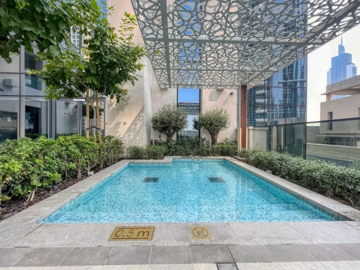 Una piscina al aire libre está bajo una cubierta calada, rodeada de vegetación y edificios modernos.