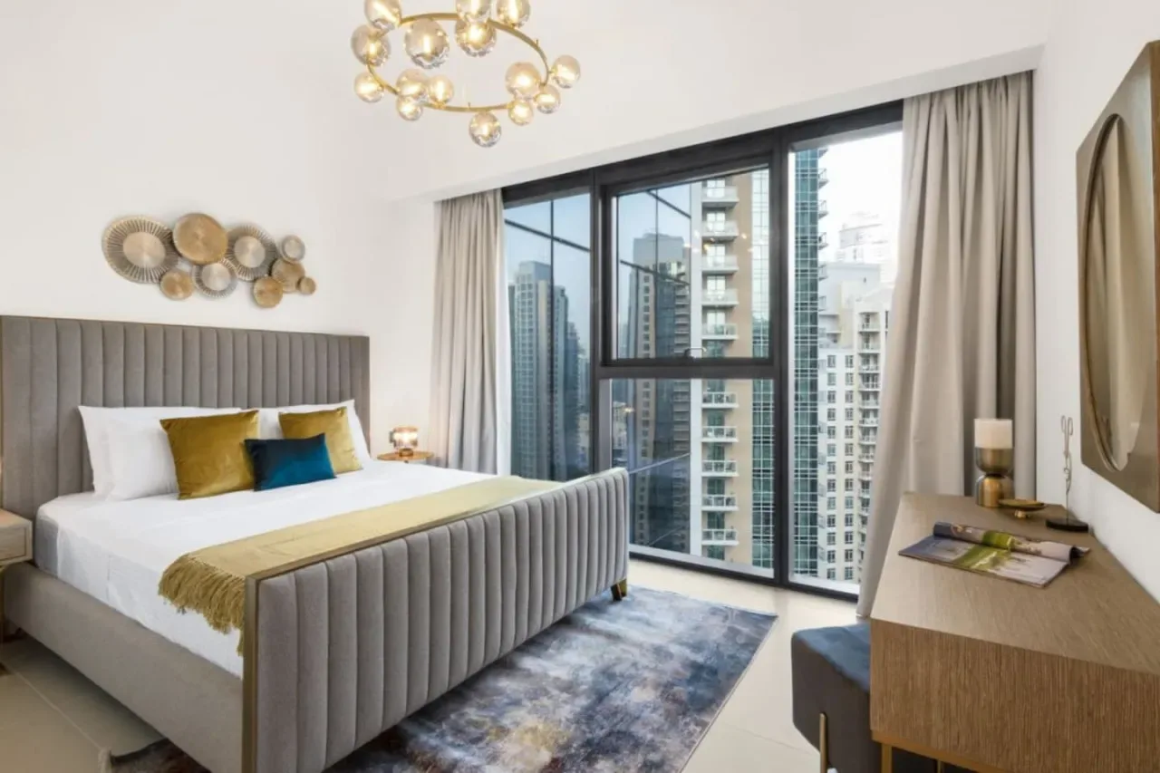 Un dormitorio con una cama grande junto a ventanales con vista a edificios cercanos.