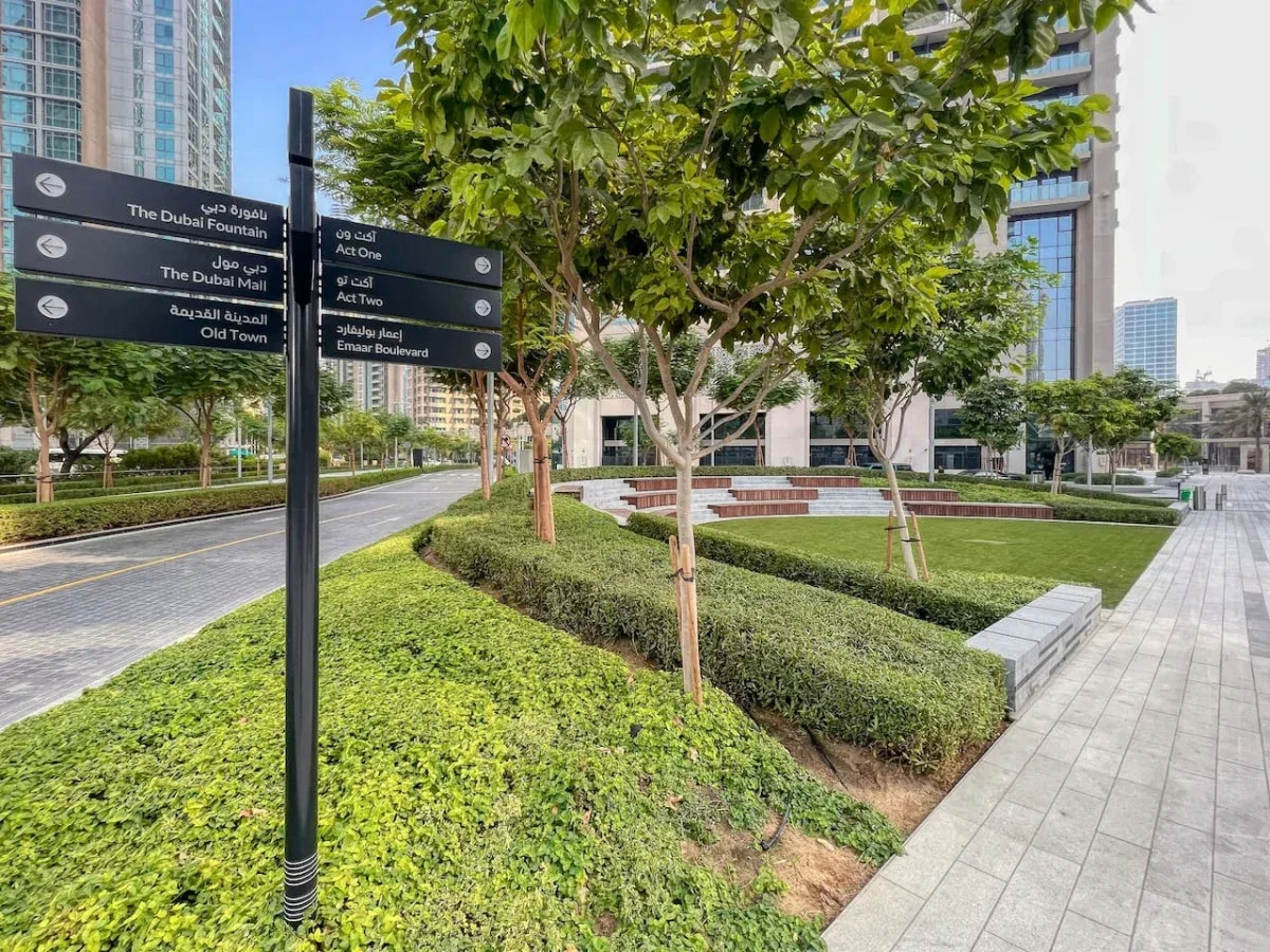 Una plaza exterior ajardinada con árboles frondosos, setos bajos y caminos pavimentados.