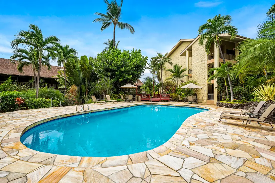 A swimming pool sits amid palm trees and low-rise buildings.