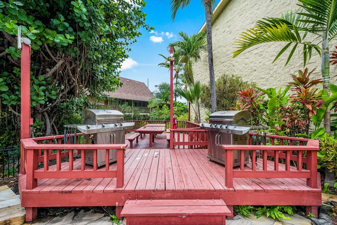 A red wooden outdoor deck with seating and two barbecue grills, surrounded by tropical greenery.