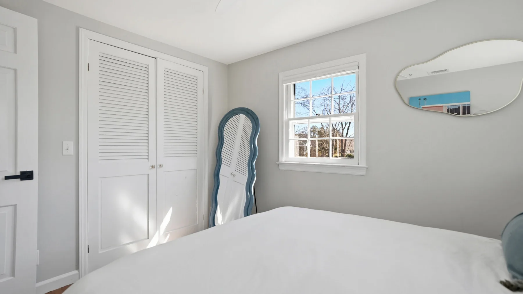 A bedroom with a bed, a window, and a tall standing mirror.