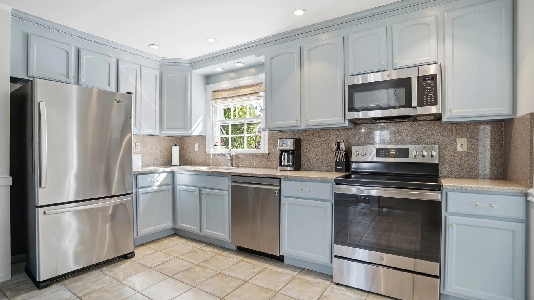 A bright kitchen with pale cabinets, stainless steel appliances, and tiled flooring.