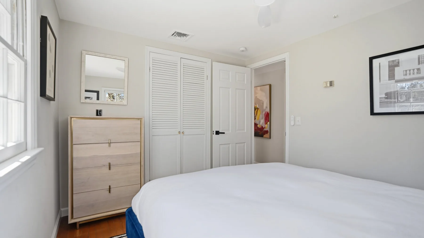 A bright bedroom with a bed, white closet doors, and a window.