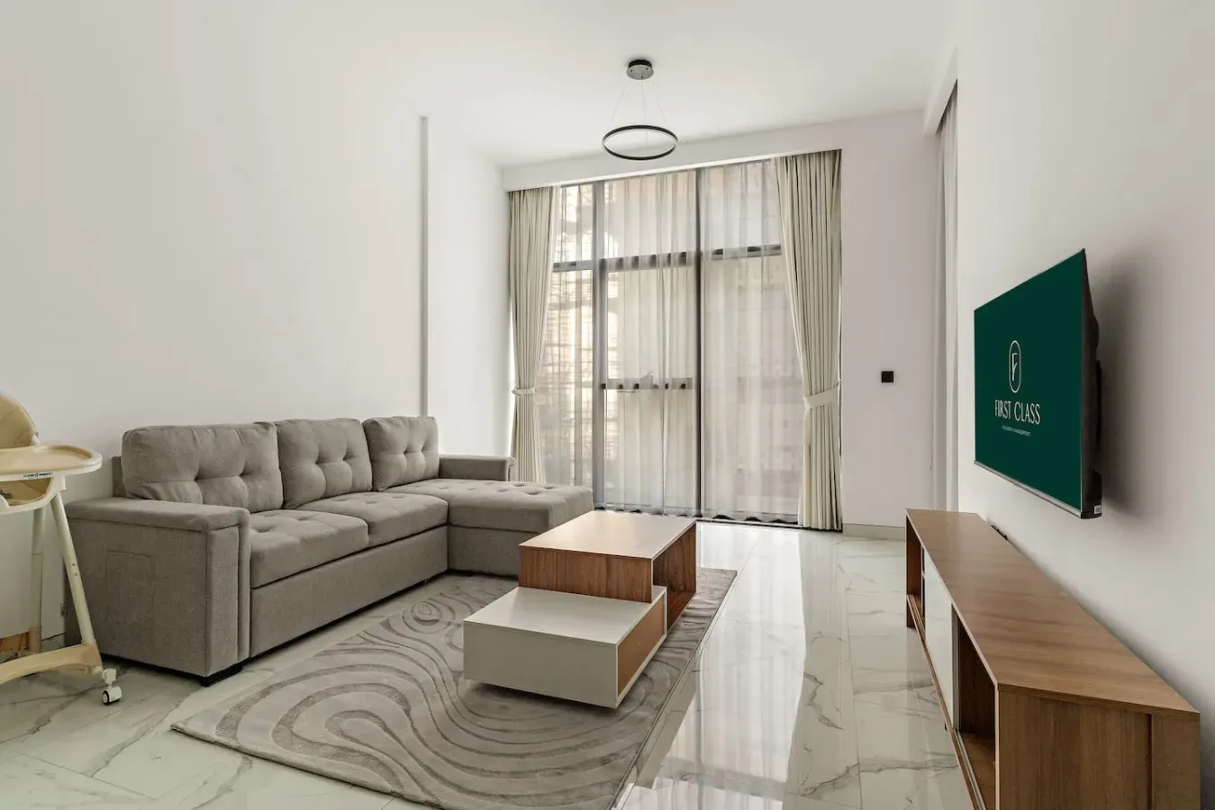 Una sala luminosa con sofá seccional gris, mesa de centro y grandes ventanas con cortinas.