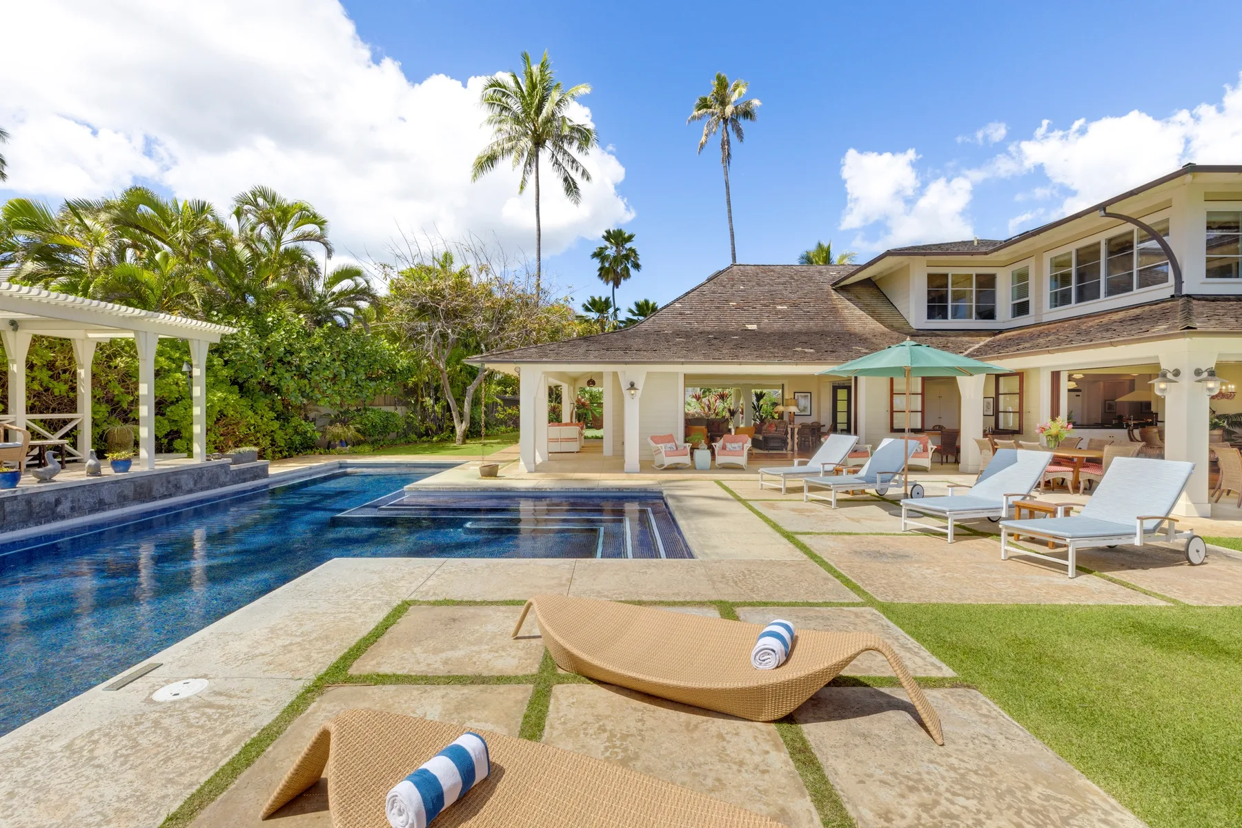 A poolside patio with lounge seating sits beside a large house and tropical landscaping.