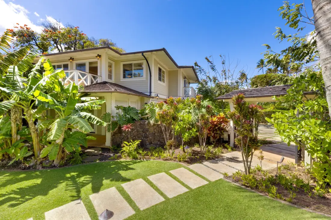 Una casa de dos plantas se encuentra detrás de un jardín tropical y un sendero de losas.