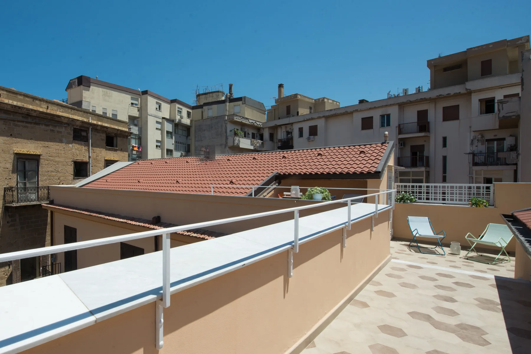 Una terraza soleada en la azotea con suelo de baldosas, tumbonas y edificios de apartamentos alrededor.