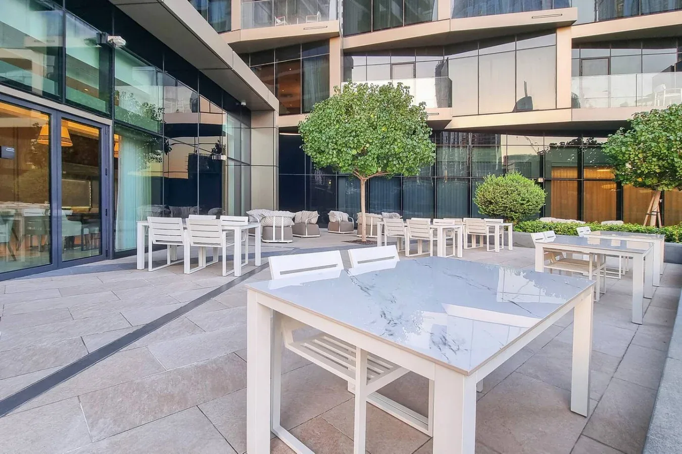 Un patio pavimentado con mesas y sillas blancas junto a un edificio moderno con fachada de vidrio.
