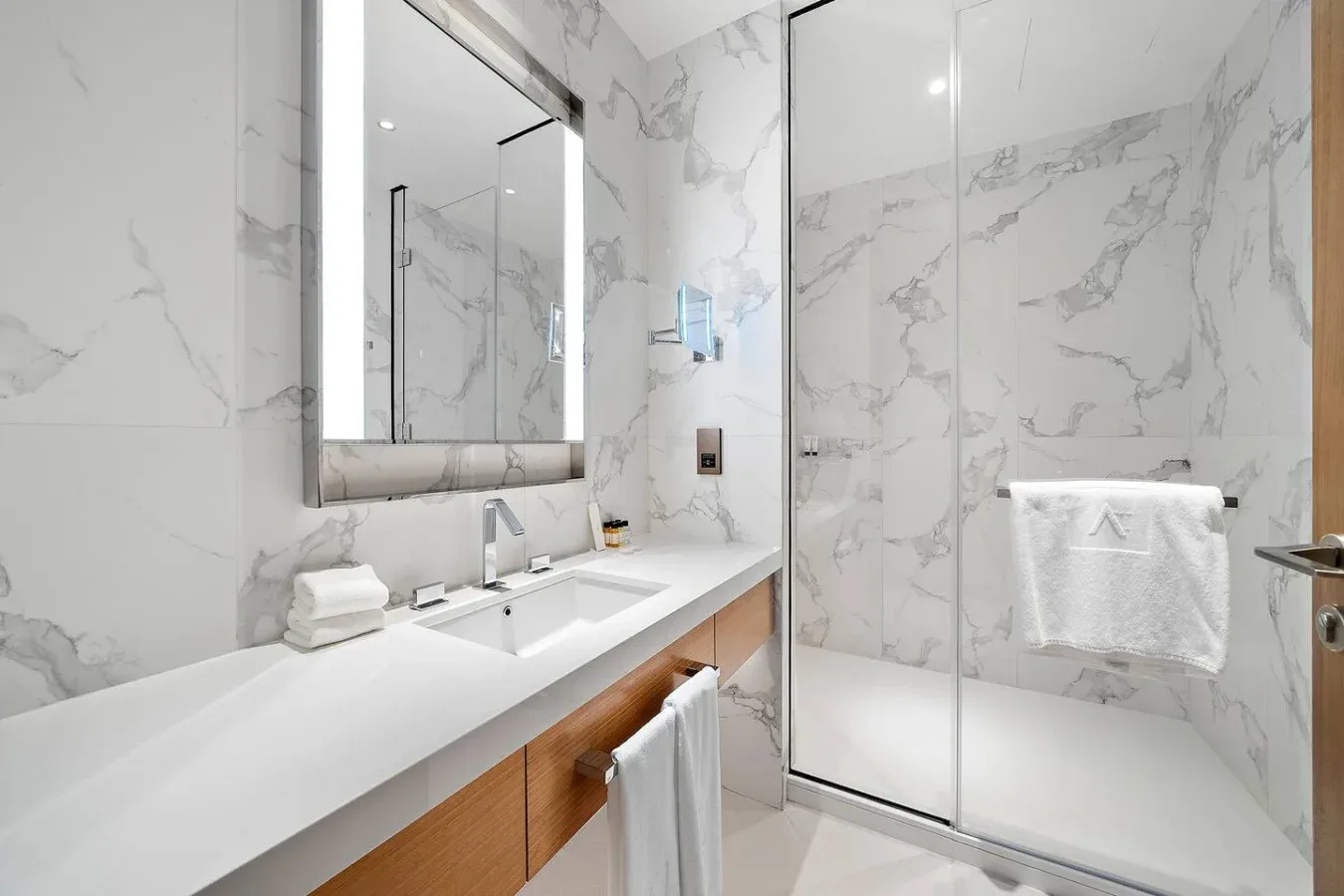 Un baño luminoso de mármol con un tocador largo, espejo y ducha de vidrio.