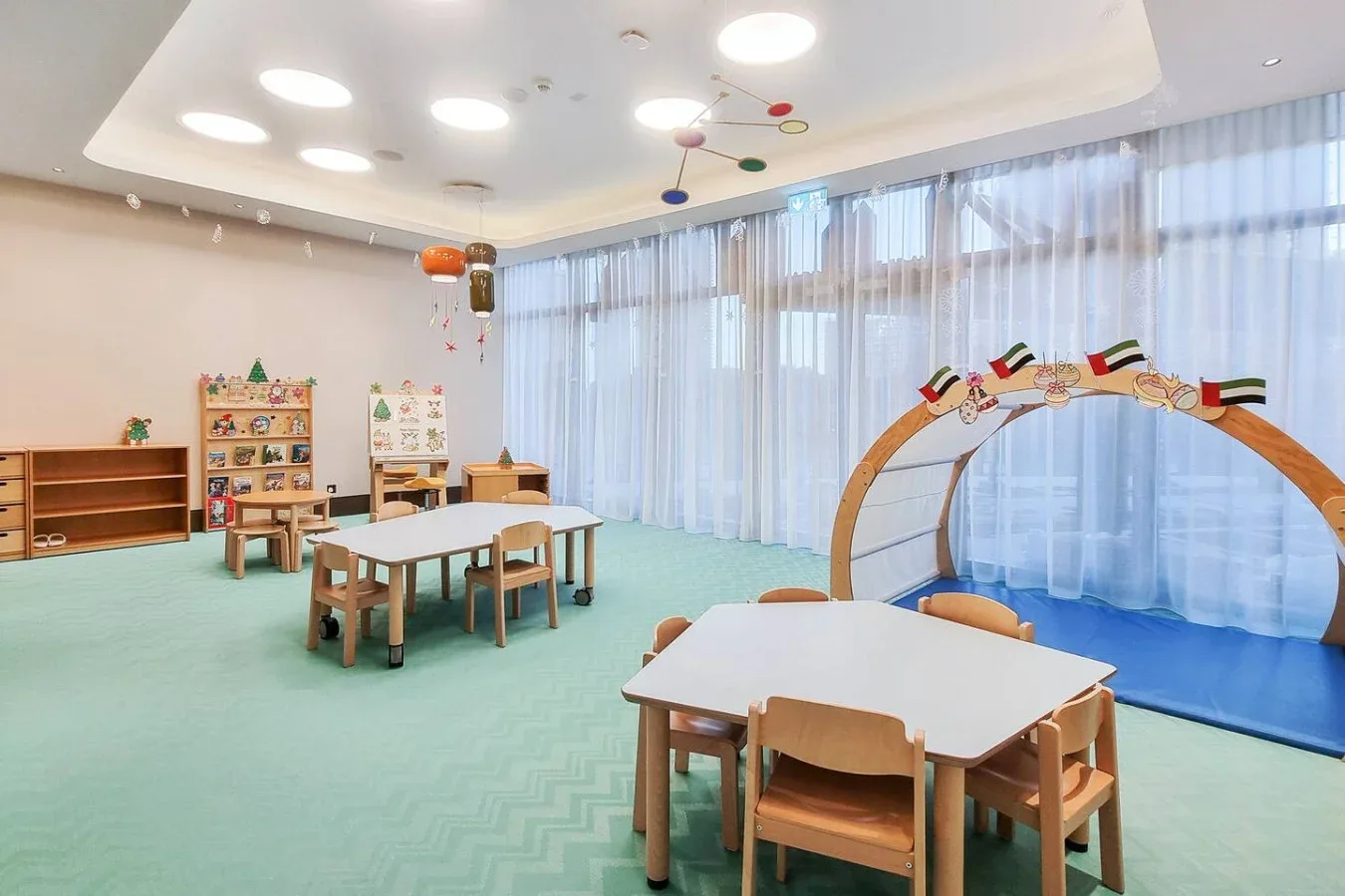 Un aula luminosa con mesas y sillas infantiles junto a grandes ventanas con cortinas.