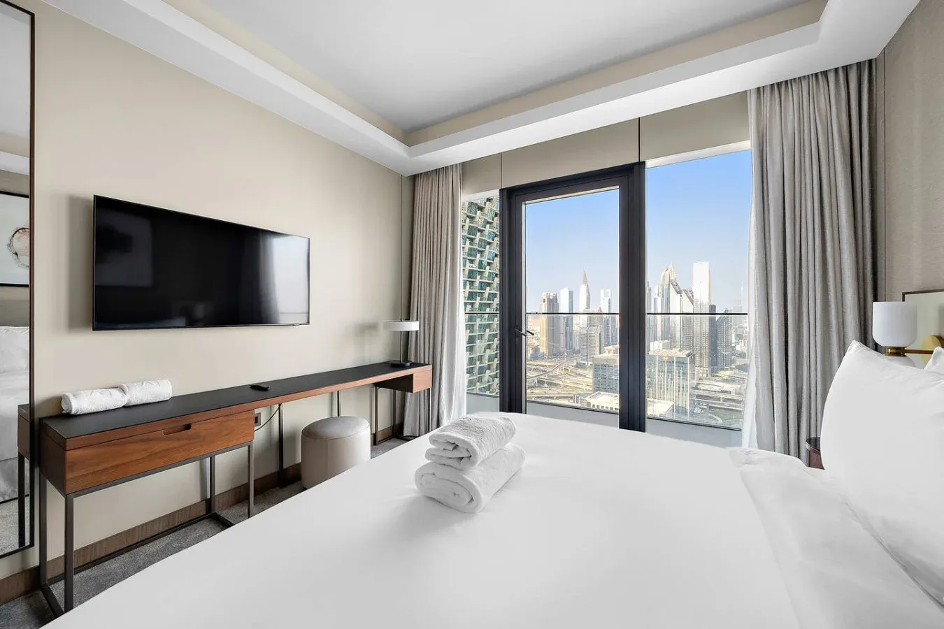 Un dormitorio con una cama blanca, un televisor en la pared y grandes ventanales con vistas a la ciudad.