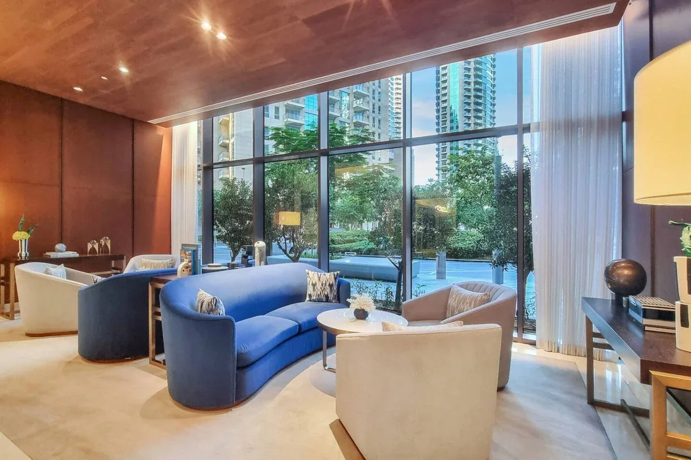 Una sala de estar con asientos azules y grandes ventanales con vistas a árboles y edificios cercanos.