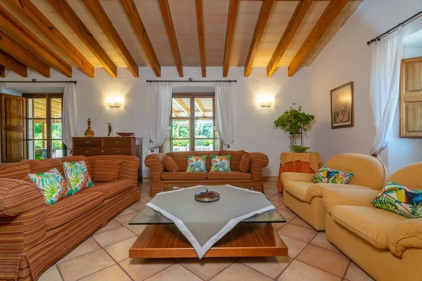 A living room with patterned seating around a low coffee table beneath exposed wooden ceiling beams.