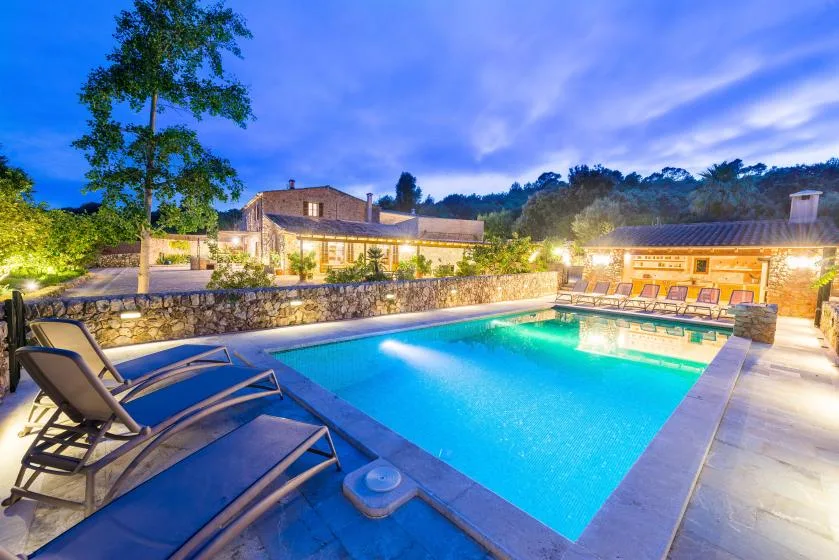 An outdoor swimming pool glows beside stone walls and a row of loungers at dusk.