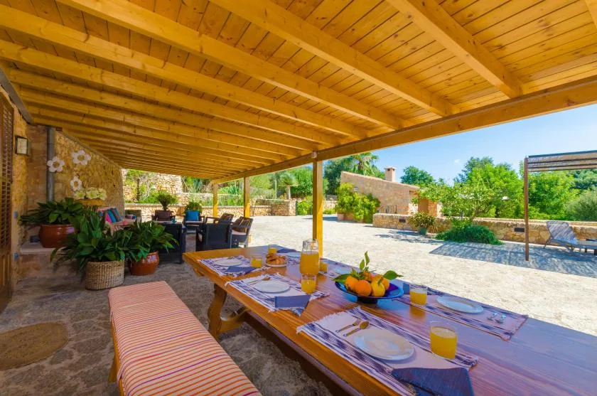 A shaded outdoor dining area with a long table beneath a wooden roof.