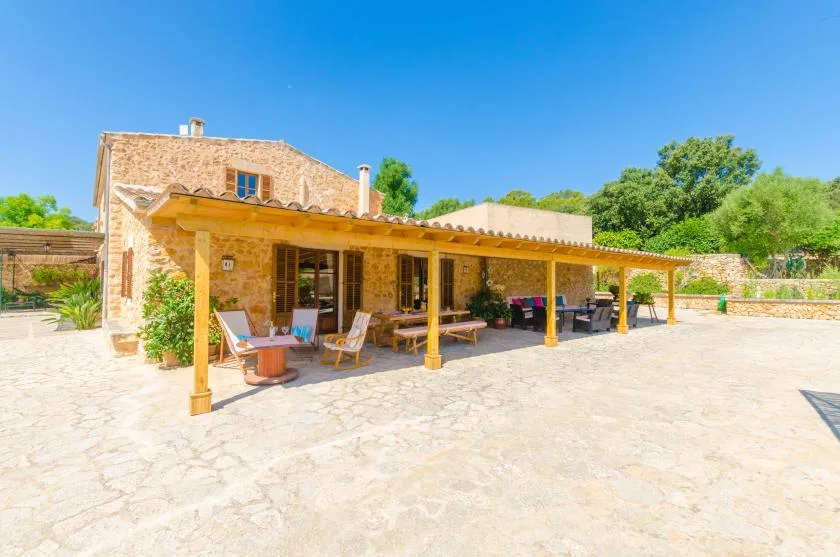 A stone house opens onto a broad paved patio shaded by a wooden pergola.