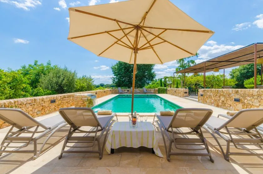 A poolside patio has lounge seating beneath a large umbrella, with greenery beyond.