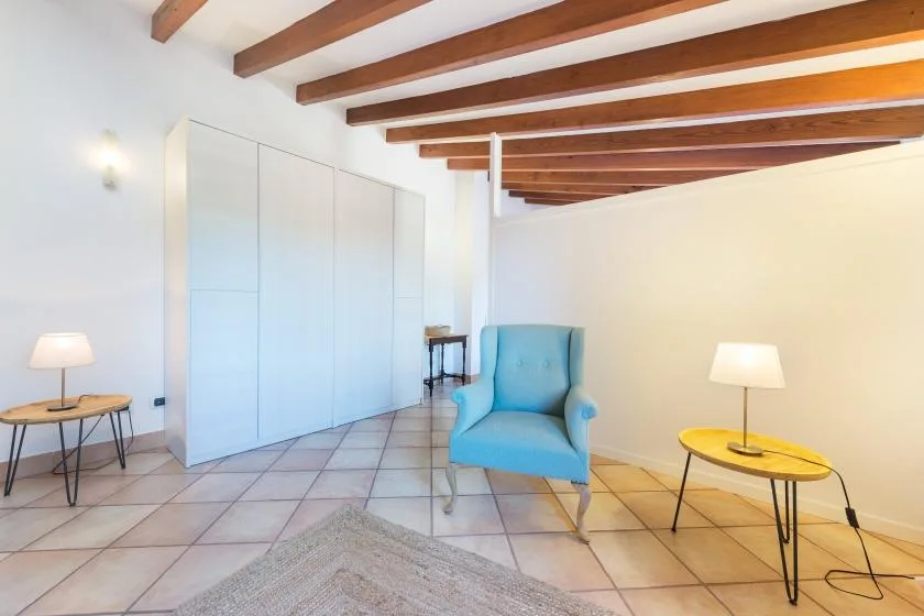 A bright sitting area with a blue armchair, tiled floor, and exposed wooden ceiling beams.