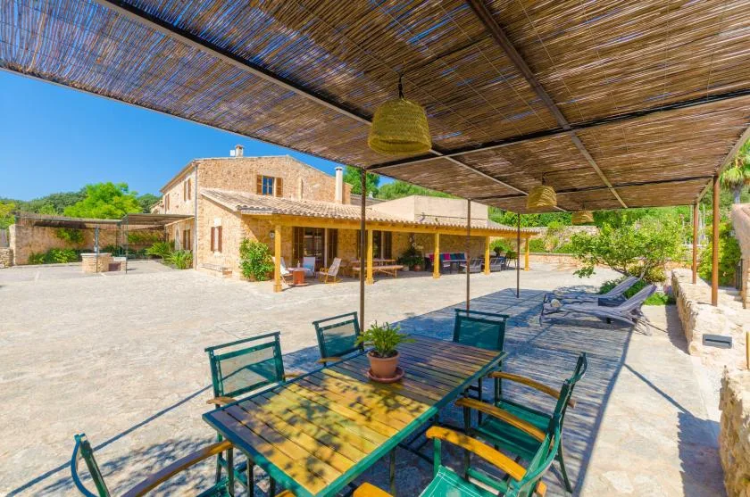 A shaded outdoor patio has a dining table and seating beside a stone building.
