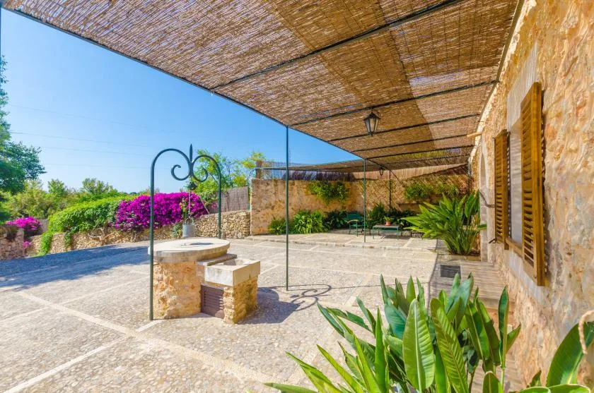 A sunlit stone patio with a woven shade canopy and surrounding plants.
