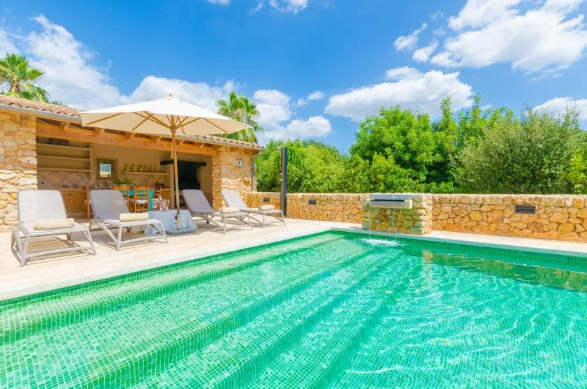 An outdoor swimming pool beside a stone patio with lounge seating and an umbrella.
