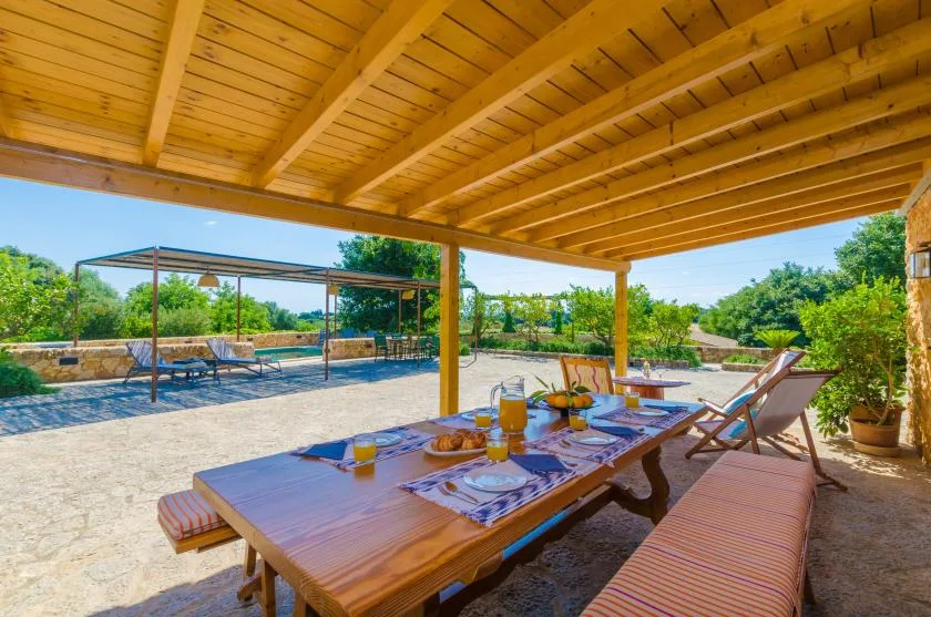 A shaded outdoor dining area beneath a wooden roof overlooks a garden.