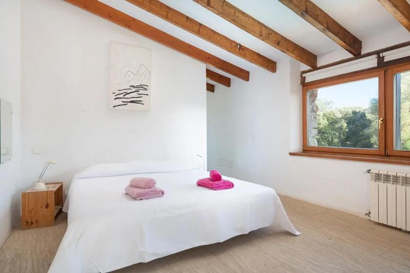 A bright bedroom with a white bed beneath exposed wooden ceiling beams.