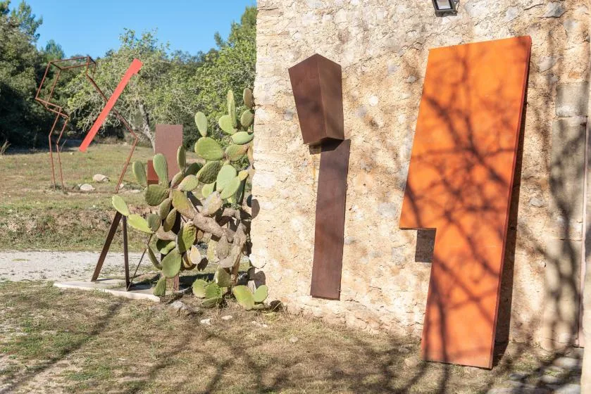 Outdoor sculpture installation beside a stone wall, with metal forms and a prickly pear cactus.