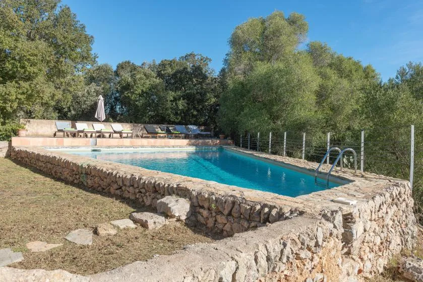 An outdoor swimming pool bordered by stone walls, with trees and seating nearby.