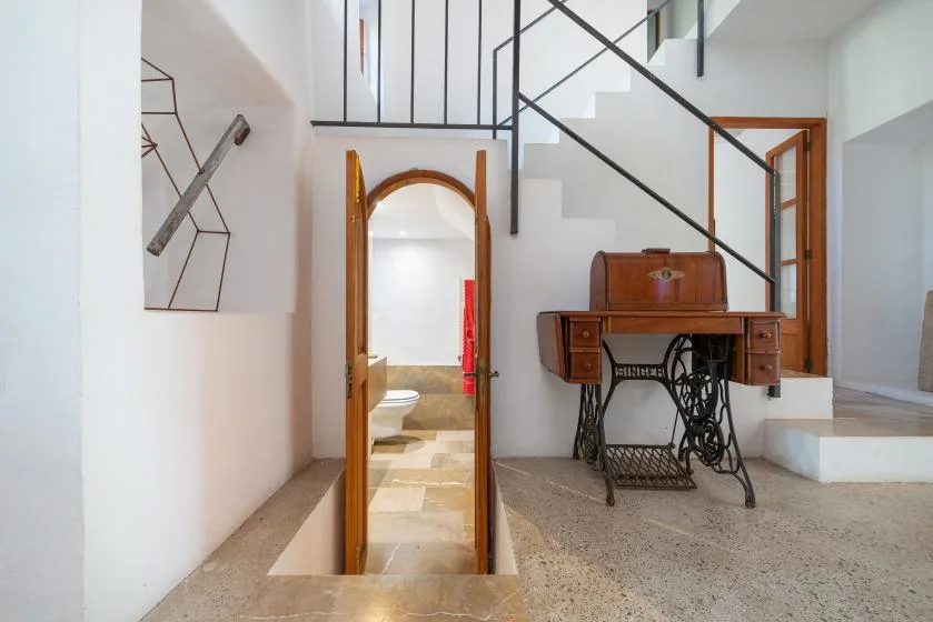A bright interior shows a staircase and an antique desk beside an arched doorway.