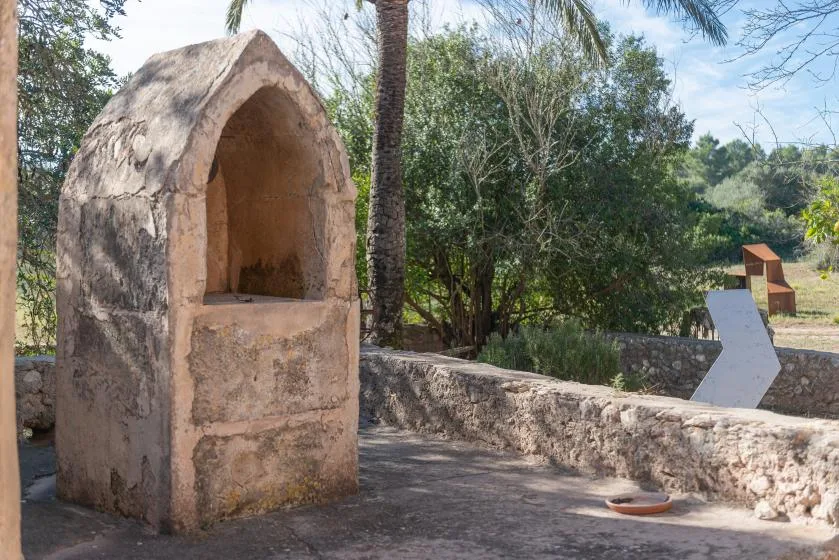 A stone outdoor structure stands beside a low wall and leafy trees.