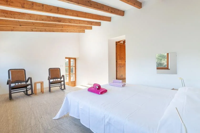 A bright bedroom with a large bed, two chairs, and exposed wooden ceiling beams.