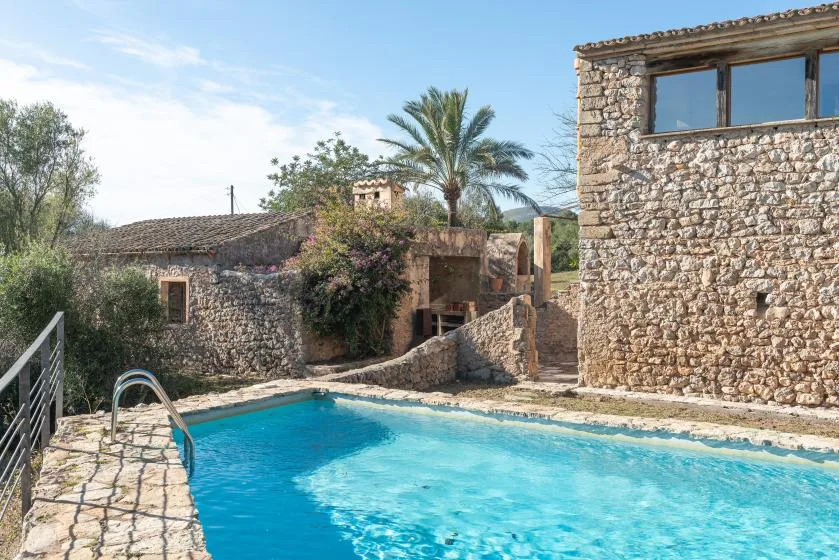 An outdoor swimming pool sits beside stone buildings and palm trees.