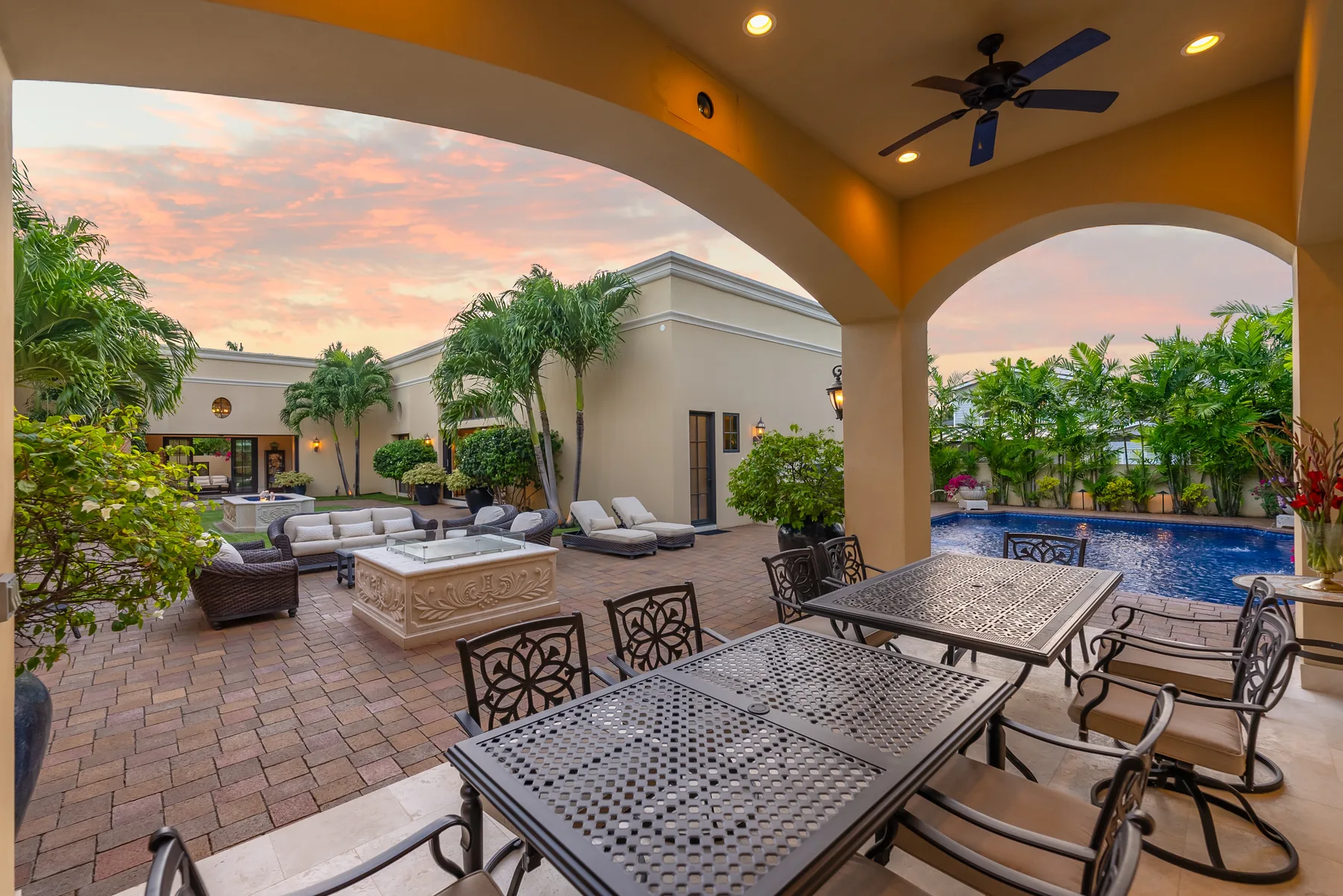Un patio exterior cubierto con mesas de comedor da a una piscina y jardines.